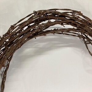 May include: A rustic, circular wreath crafted from weathered, rusty barbed wire. The brown metal wire is tightly wound, creating a textured, semi-circular shape. The wreath is set against a plain white background, highlighting its rough, industrial aesthetic.