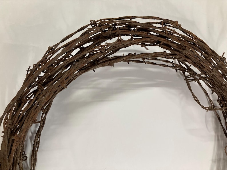 May include: A rustic, circular wreath crafted from weathered, rusty barbed wire. The brown metal wire is tightly wound, creating a textured, semi-circular shape. The wreath is set against a plain white background, highlighting its rough, industrial aesthetic.