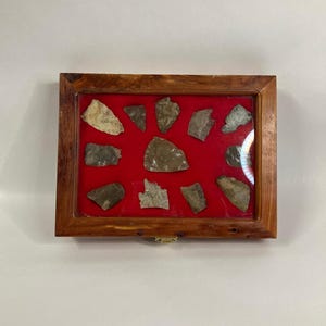May include: A wooden display case with a red velvet interior, showcasing a collection of 12 arrowheads.