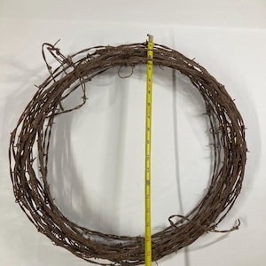 May include: A circular wreath made of rusty barbed wire. The wreath is approximately 10 inches in diameter, as measured by a yellow tape measure. The barbed wire is a dark brown color.