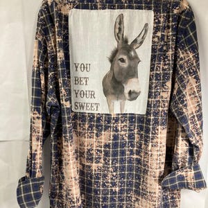 May include: A blue and white plaid flannel shirt with a bleached effect. The back of the shirt features a patch with a donkey and the text "You Bet Your Sweet".