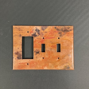 May include: A copper-colored wall plate with a single light switch and two toggle switches. The plate has a rustic, distressed finish.