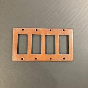 May include: A copper-colored metal plate with four rectangular cutouts for light switch plates. The plate has a hammered texture and eight small holes around the edges.
