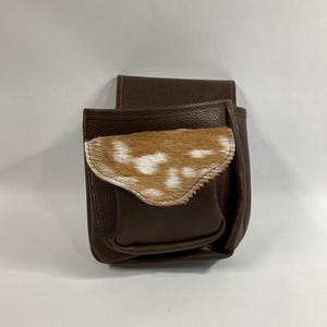 May include: A brown leather pouch with a flap that has a white and brown spotted fur lining. The pouch has a zipper closure.
