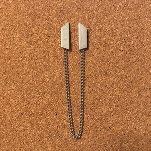 May include: Two silver metal pieces with a chain connecting them. The pieces are shaped like a triangle with a rounded edge. The chain is a simple link chain.
