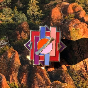 Peut inclure: Une épingle en émail dorée avec un motif géométrique. L'épingle présente un schéma de couleurs rouge, rose, orange et bleu. L'épingle est placée sur un fond de rochers et d'arbres.