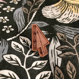 May include: Wooden arrow-shaped pin with the word "BOOP" written vertically on the front. The pin is on a dark green fabric with a floral pattern.