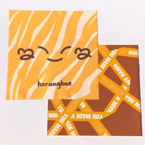 May include: A yellow microfiber cleaning cloth with a tiger stripe pattern and a cartoon smiley face. The text "horanghae" is printed on the cloth. The cloth is folded in half and is sitting on top of a brown and yellow patterned fabric.