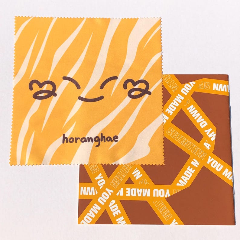May include: A yellow microfiber cleaning cloth with a tiger stripe pattern and a cartoon smiley face. The text "horanghae" is printed on the cloth. The cloth is folded in half and is sitting on top of a brown and yellow patterned fabric.