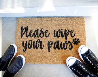 May include: A brown coir doormat with the text "Please wipe your paws" in black lettering and a paw print icon. Two pairs of black shoes are on either side of the doormat.