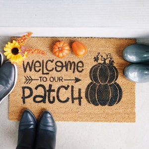 May include: A brown coir doormat with the text "Welcome to our Patch" in black lettering. The doormat features a black silhouette of two pumpkins. There are two orange pumpkins and a bouquet of yellow flowers on the doormat.