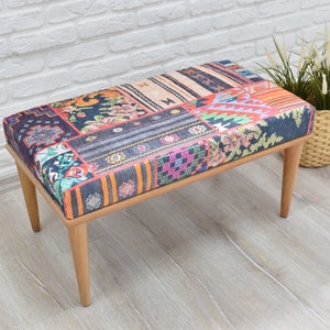 Pode incluir: Banqueta retangular com assento estampado em patchwork multicolorido, incluindo azul, laranja e rosa. A banqueta tem quatro pernas de madeira. &Agrave; direita, uma cesta de vime com plantas. A banqueta est&aacute; em frente a uma parede de tijolos brancos.