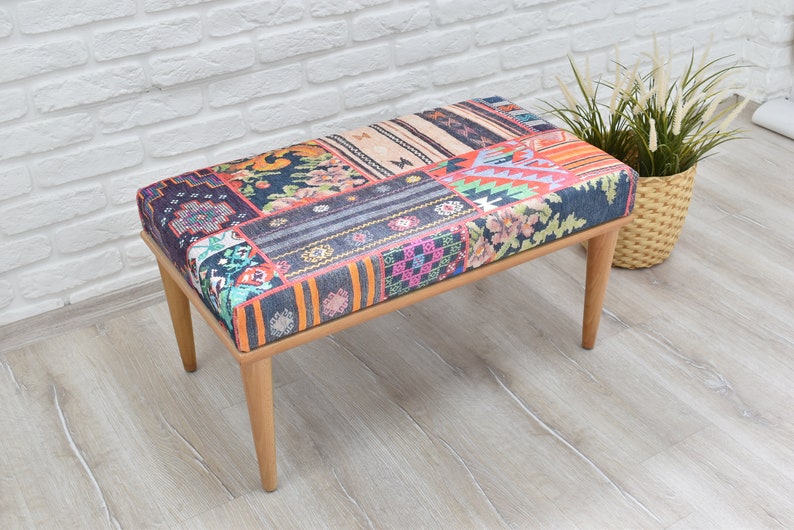 Pode incluir: Banqueta retangular com assento estampado em patchwork multicolorido, incluindo azul, laranja e rosa. A banqueta tem quatro pernas de madeira. &Agrave; direita, uma cesta de vime com plantas. A banqueta est&aacute; em frente a uma parede de tijolos brancos.