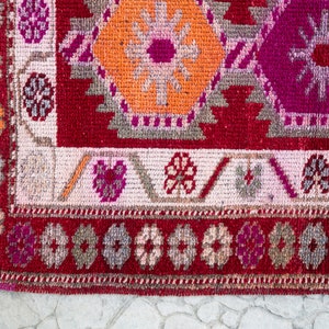 May include: Close-up of a red, pink, and orange patterned rug with a fringed edge. The rug has a geometric design with floral motifs.