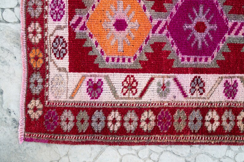 May include: Close-up of a red, pink, and orange patterned rug with a fringed edge. The rug has a geometric design with floral motifs.