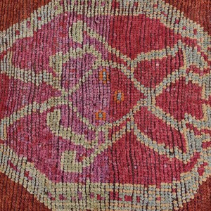 May include: Close-up of a textured, handmade rug with a repeating geometric pattern. The rug features a central diamond shape in shades of red and pink, surrounded by a border of gray and orange. The rug's design is intricate and detailed.