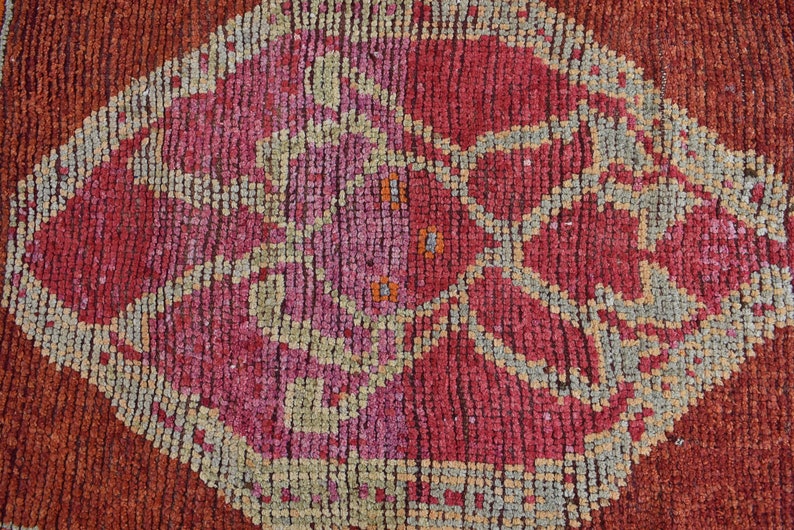 May include: Close-up of a textured, handmade rug with a repeating geometric pattern. The rug features a central diamond shape in shades of red and pink, surrounded by a border of gray and orange. The rug's design is intricate and detailed.