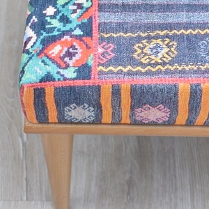 Pode incluir: Close-up de um pufe estampado com estrutura de madeira e pernas afiladas. O tecido apresenta um design de patchwork com v&aacute;rias cores, incluindo azul, laranja, rosa e verde. As pernas s&atilde;o castanho claro.