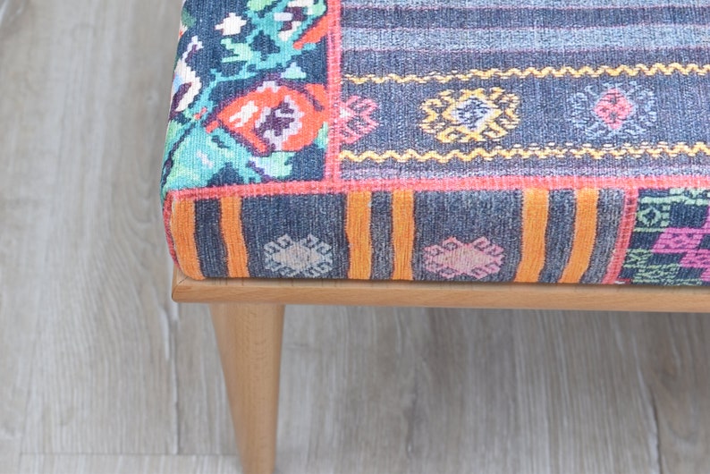 Pode incluir: Close-up de um pufe estampado com estrutura de madeira e pernas afiladas. O tecido apresenta um design de patchwork com v&aacute;rias cores, incluindo azul, laranja, rosa e verde. As pernas s&atilde;o castanho claro.