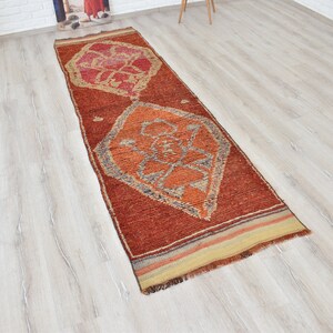 May include: A rectangular, rust-colored runner rug with geometric designs in orange, red, and gray. The rug features a central diamond pattern and a striped border. The rug is placed on a light wood floor.