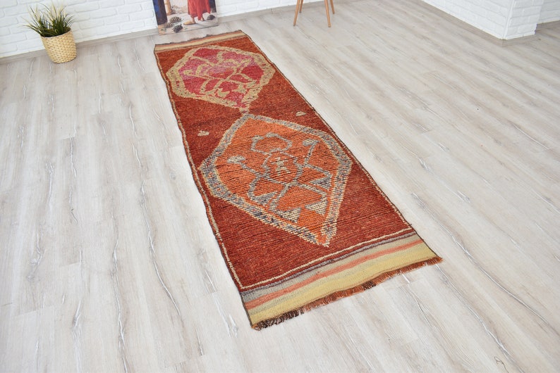 May include: A rectangular, rust-colored runner rug with geometric designs in orange, red, and gray. The rug features a central diamond pattern and a striped border. The rug is placed on a light wood floor.