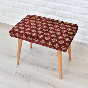 May include: A small, red upholstered stool with a gold patterned fabric. The stool has four wooden legs.