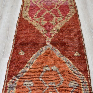 May include: A rectangular, hand-woven rug with a geometric design. The rug features a central pattern in shades of orange, red, and pink, with a darker red border. The design includes diamond and triangle shapes. The rug is placed on a light wood floor.