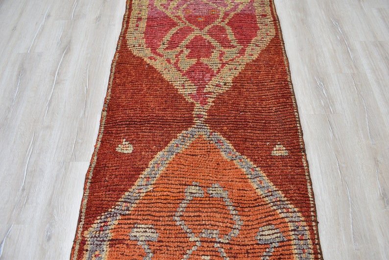 May include: A rectangular, hand-woven rug with a geometric design. The rug features a central pattern in shades of orange, red, and pink, with a darker red border. The design includes diamond and triangle shapes. The rug is placed on a light wood floor.