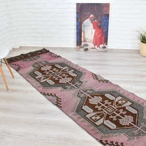 May include: A pink and gray patterned runner rug with a geometric design. The rug has a traditional style and is made of wool.