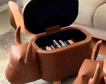 May include: A brown leather ottoman shaped like a bull with a hinged lid that opens to reveal storage space. The ottoman is sitting on a white rug and has wooden legs.