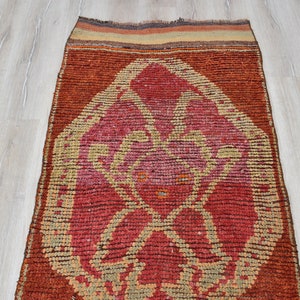May include: A rectangular, hand-woven rug with a central geometric design in shades of red, pink, and beige. The rug has a dark brown border and a textured appearance. The rug is approximately 60 inches long.