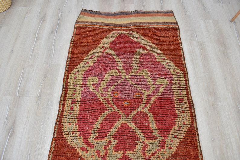 May include: A rectangular, hand-woven rug with a central geometric design in shades of red, pink, and beige. The rug has a dark brown border and a textured appearance. The rug is approximately 60 inches long.
