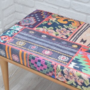 Pode incluir: Banqueta retangular com design de patchwork. O assento apresenta uma variedade de padr&otilde;es coloridos, incluindo motivos florais, geom&eacute;tricos e listrados. A banqueta tem pernas de madeira de cor clara.