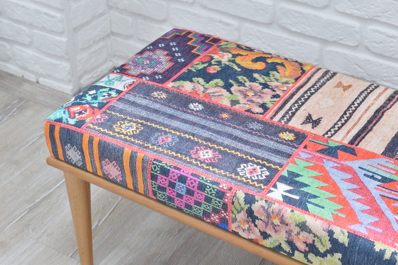 Pode incluir: Banqueta retangular com design de patchwork. O assento apresenta uma variedade de padr&otilde;es coloridos, incluindo motivos florais, geom&eacute;tricos e listrados. A banqueta tem pernas de madeira de cor clara.