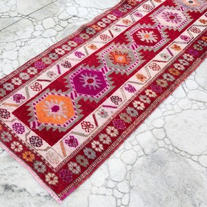May include: A red, pink, and orange patterned runner rug with a geometric design. The rug has a fringe border.