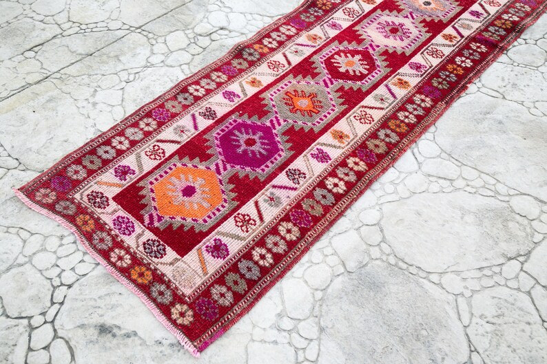 May include: A red, pink, and orange patterned runner rug with a geometric design. The rug has a fringe border.