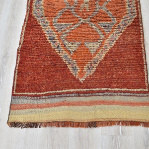 May include: A rectangular, hand-woven rug with a geometric diamond pattern in shades of orange, beige, and gray. The rug has a textured appearance and a fringe along the bottom edge. The rug is placed on a light-colored wooden floor.