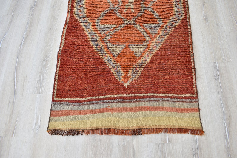 May include: A rectangular, hand-woven rug with a geometric diamond pattern in shades of orange, beige, and gray. The rug has a textured appearance and a fringe along the bottom edge. The rug is placed on a light-colored wooden floor.