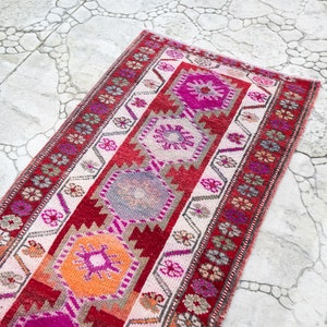 May include: A long, narrow rug with a traditional Turkish design. The rug is made of wool and has a cream background with red, pink, orange, and gray geometric patterns.