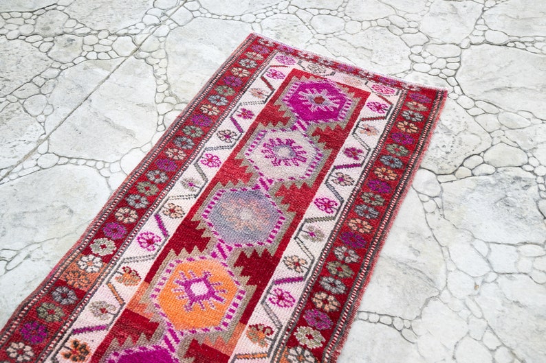 May include: A long, narrow rug with a traditional Turkish design. The rug is made of wool and has a cream background with red, pink, orange, and gray geometric patterns.