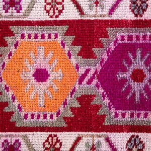 May include: Close-up of a colorful rug with a geometric pattern. The rug features a red background with orange, pink, and gray accents. The pattern is made up of repeating shapes and lines.