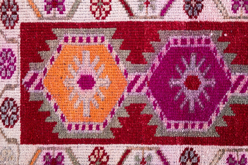 May include: Close-up of a colorful rug with a geometric pattern. The rug features a red background with orange, pink, and gray accents. The pattern is made up of repeating shapes and lines.