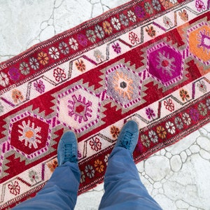 May include: A red, pink, and orange patterned rug with a geometric design. The rug is laid out on a white tiled floor. A person's feet are visible in the bottom of the image, wearing blue jeans and blue shoes.