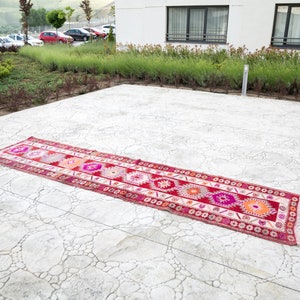 May include: A long, narrow, red and white patterned rug with geometric designs lies on a gray concrete patio.