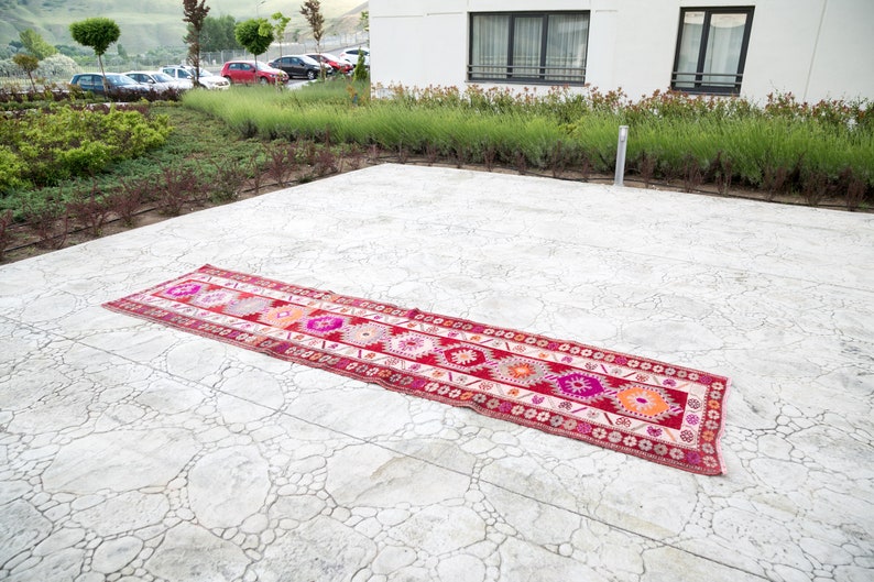 May include: A long, narrow, red and white patterned rug with geometric designs lies on a gray concrete patio.
