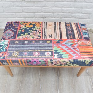 Pode incluir: Um pufe retangular com um design de patchwork. A parte superior apresenta v&aacute;rios padr&otilde;es de tecido em tons de azul, laranja, rosa e bege. Possui pernas de madeira de cor clara e est&aacute; colocado sobre um piso de madeira clara.