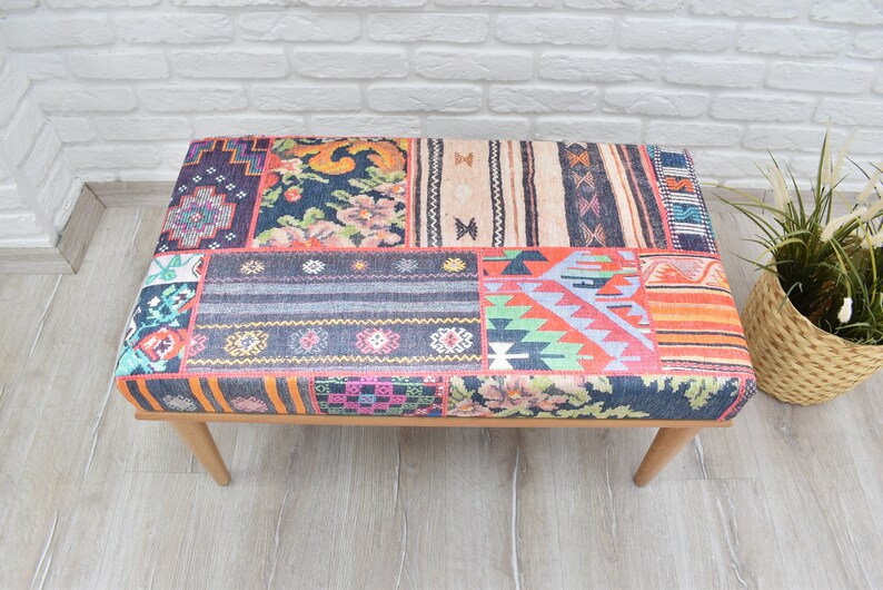 Pode incluir: Um pufe retangular com um design de patchwork. A parte superior apresenta v&aacute;rios padr&otilde;es de tecido em tons de azul, laranja, rosa e bege. Possui pernas de madeira de cor clara e est&aacute; colocado sobre um piso de madeira clara.