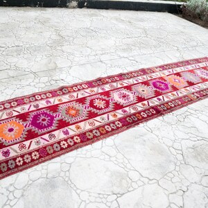 May include: A long, narrow red, pink, and white patterned rug with a geometric design. The rug is laid out on a cracked concrete surface.