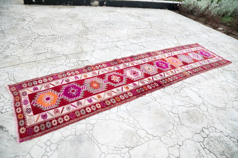 May include: A long, narrow red, pink, and white patterned rug with a geometric design. The rug is laid out on a cracked concrete surface.