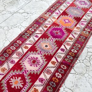 May include: A long, narrow red and white runner rug with a geometric pattern in shades of pink, orange, and gray. The rug has a traditional Turkish design.